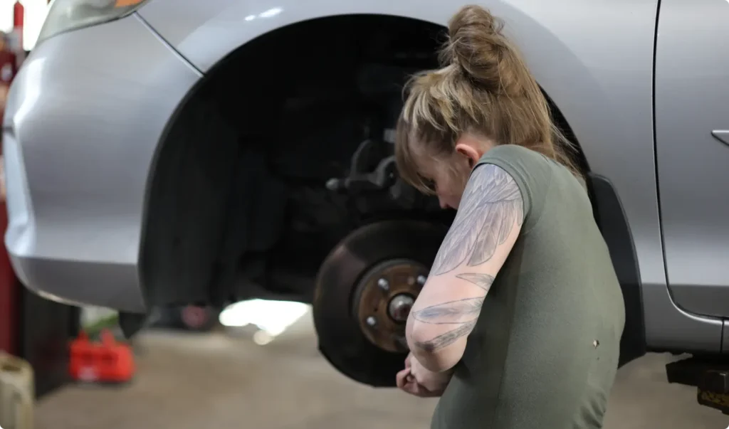 A woman with a tattooed arm works on repairing a car with a missing wheel in a garage. The cars undercarriage and brake disc are visible as she focuses on her task.