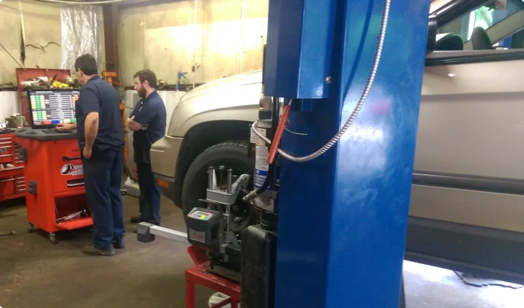 Two mechanics stand near a red tool chest in an auto repair shop. Part of a beige SUV is lifted on a hydraulic car lift, with various tools and equipment visible around the workshop.