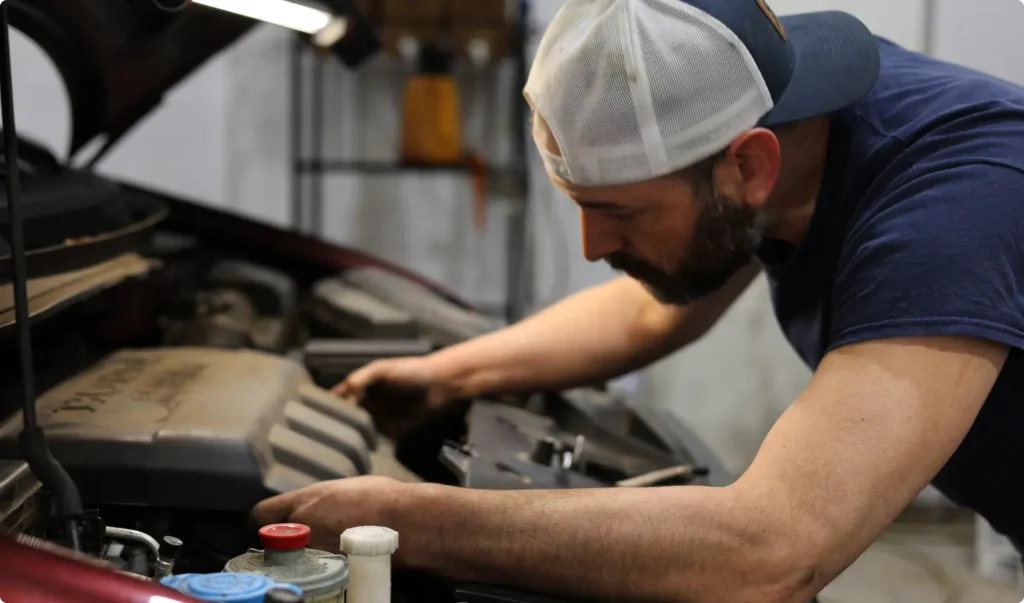 A man wearing a white baseball cap and navy shirt inspects or repairs the engine of a car with the hood open, focusing intently while working in a garage or workshop.
