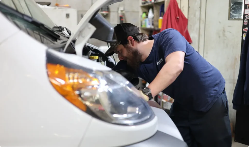 A mechanic wearing a navy shirt and black cap works under the hood of a white vehicle in a garage, focusing on the engine area with tools and shelves visible in the background.
