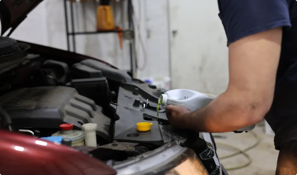A person is pouring engine oil into a cars engine while the hood is open, in what appears to be a garage or workshop setting.
