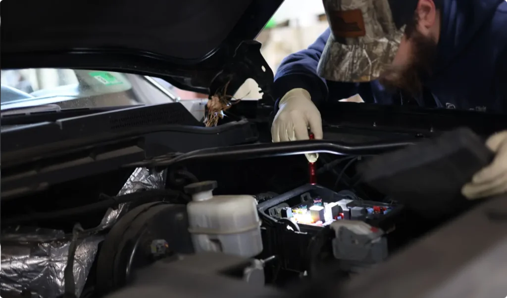 A person wearing gloves and a camouflage hat works under the hood of a car, using a tool to inspect or repair the engine components. The vehicles engine bay is well-lit, showing various parts and fluid containers.