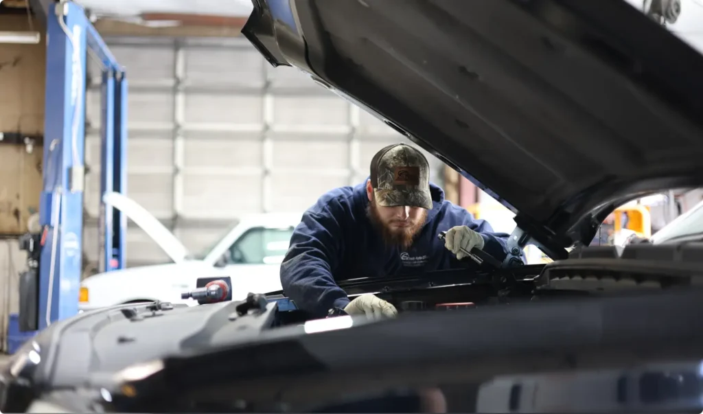 A mechanic wearing a cap and gloves works intently under the hood of a car in a garage, with another white vehicle visible in the background.