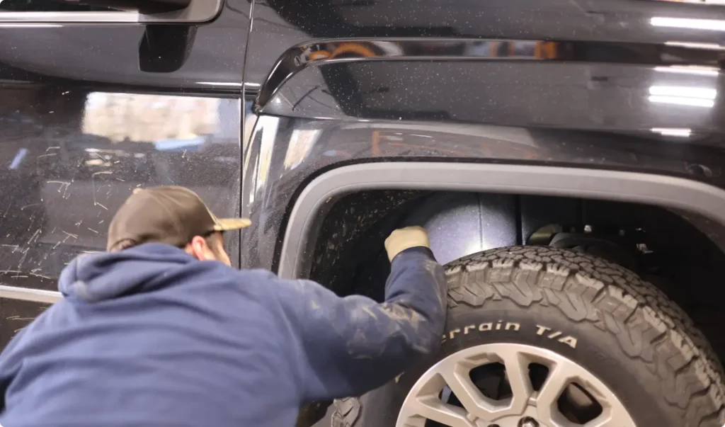 A person wearing a cap and gloves inspects or works on the wheel well of a black SUV, focusing on the area above the tire.