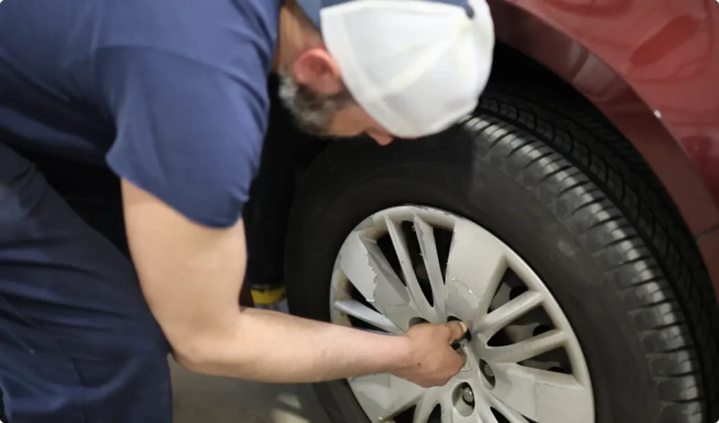 A person wearing a white cap and blue shirt uses a tool to loosen or tighten the lug nuts on the wheel of a red car, possibly performing maintenance or changing a tire.