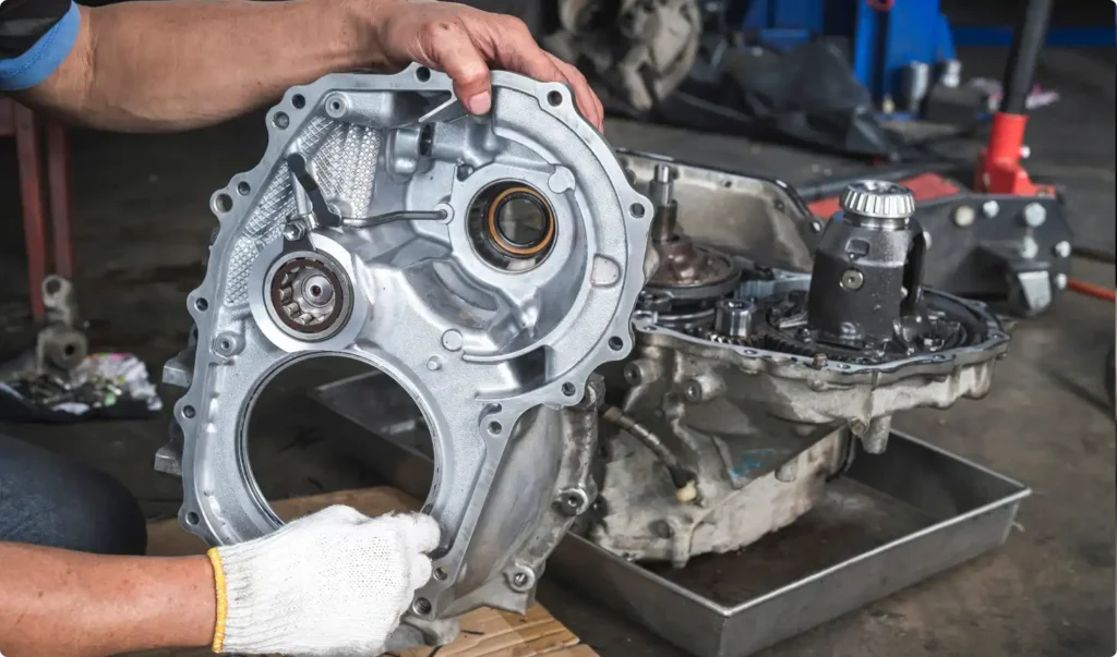 A person wearing gloves holds a metallic car transmission case cover near an exposed transmission assembly in an auto repair workshop.