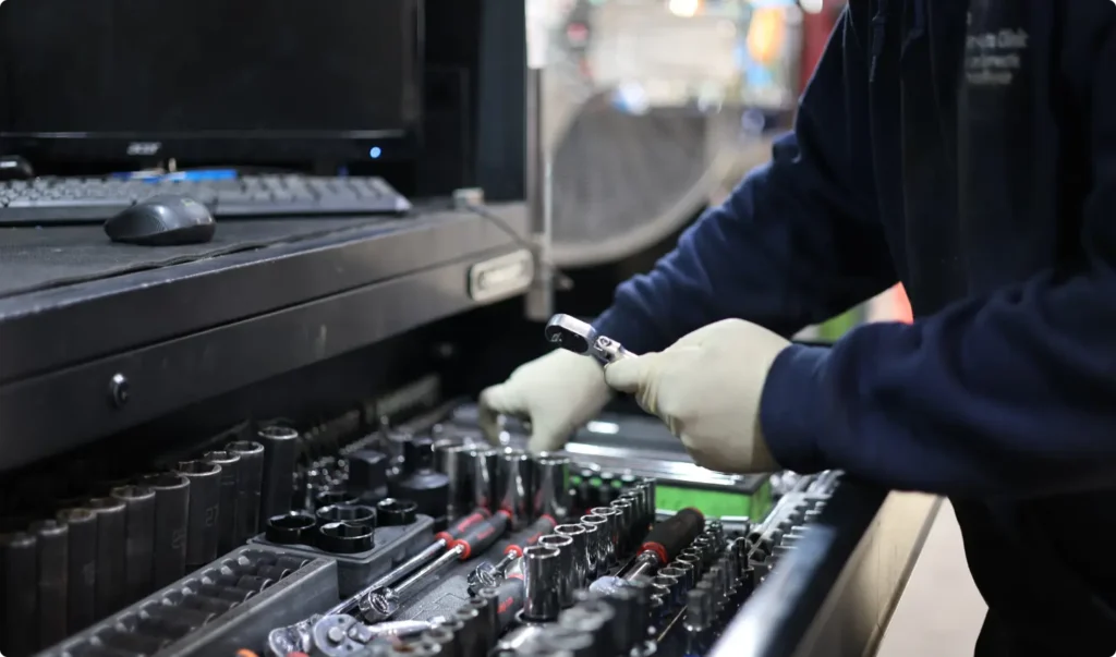 A person wearing gloves is selecting a tool from an organized drawer filled with various hand tools and sockets, with a computer keyboard visible on the workstation above.