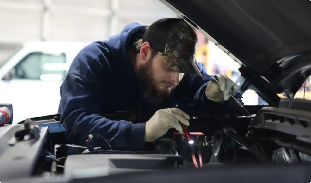 A mechanic wearing gloves, a blue hoodie, and a camouflage cap is working intently under the hood of a vehicle, holding a tool and inspecting the engine in a garage setting.