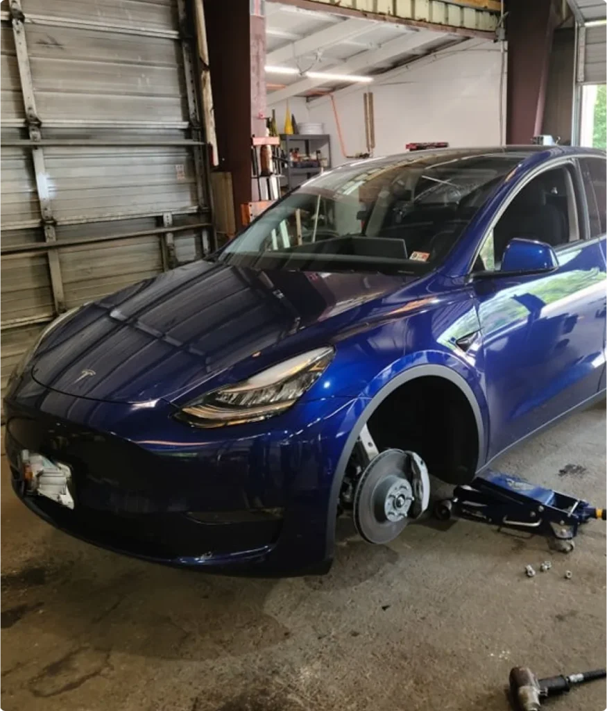 A blue Tesla car inside a garage with its front left wheel removed, sitting on a jack. Tools and lug nuts are scattered on the floor nearby.