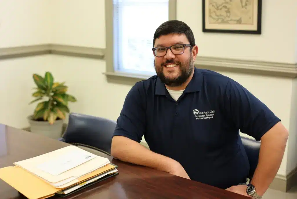 A smiling man with glasses and a beard sits at a desk with paperwork. He wears a navy polo shirt with a logo. Behind him is a window, a potted plant, and a framed map on the wall.