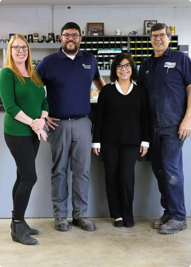 Four adults stand side by side, smiling, in front of a workbench with small shelves and organized containers behind them. Two are dressed casually, one in business attire, and one in work clothes.