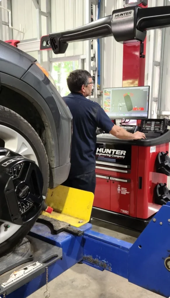 A mechanic stands at a computer workstation using a Hunter alignment machine, with a car mounted on a blue hydraulic lift in an auto repair shop. The computer screen displays alignment data for the vehicle.