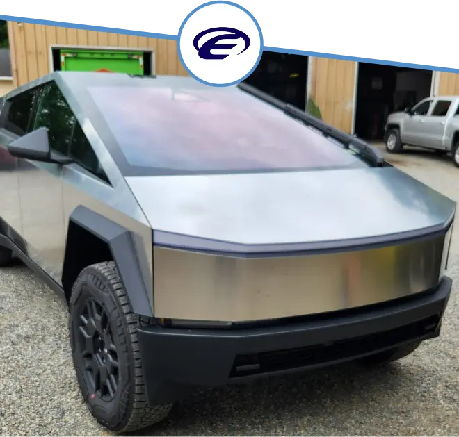 A silver Tesla Cybertruck with black wheels is parked on a paved surface in front of a building with wooden siding. There is a logo featuring a stylized E above the truck.