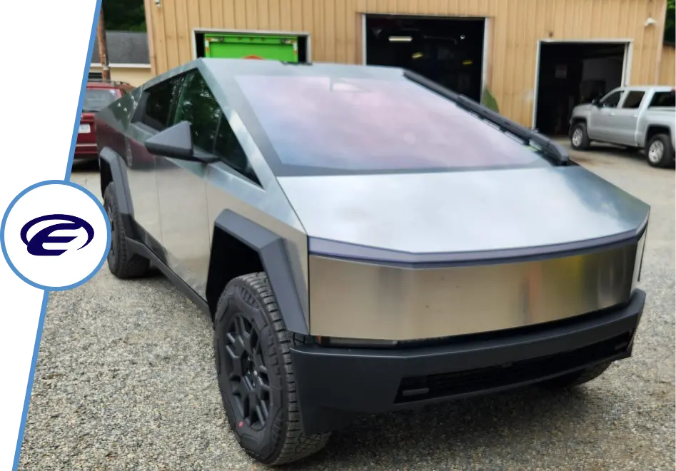 A silver Tesla Cybertruck is parked on a gravel lot in front of a beige building, with other vehicles and a green garage door in the background. A stylized E logo is visible on the left side of the image.