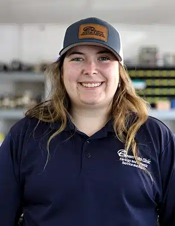 Haley Oliver, a Service Advisor, smiles at the camera. She is wearing a dark blue polo shirt with 'Elham Auto Clinic' embroidered on it and a baseball cap with a patch that also says 'Elham Auto Clinic'.