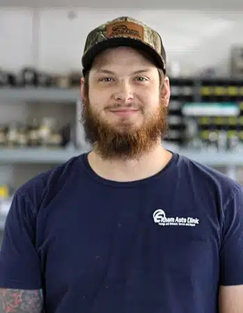 Kevn Massey, a Technician at Elham Auto Clinic, smiles at the camera. He has a beard and is wearing a dark blue polo shirt with the 'Elham Auto Clinic' logo and a camouflage baseball cap.