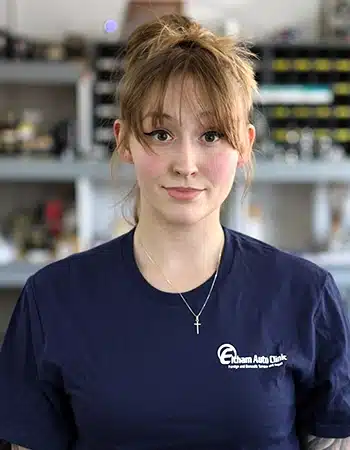 Marissa Halbig, a Technician at Elham Auto Clinic, looks directly at the camera with a subtle smile. She has light brown hair tied up, bangs, and is wearing a dark blue polo shirt with the 'Elham Auto Clinic' logo and a silver cross necklace.