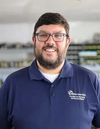 our team, auto repair in West Point, VA at Eltham Auto Clinic. Travis Moskalski, Owner/President of Elham Auto Clinic, smiles at the camera. He has a beard, is wearing glasses, and a dark blue polo shirt with the 'Elham Auto Clinic' logo.