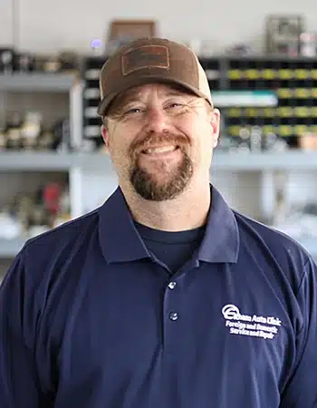 David Dean, the Lead Technician at Elham Auto Clinic, smiles broadly at the camera. He has a goatee, is wearing a dark blue polo shirt with the 'Elham Auto Clinic' logo, and a brown baseball cap.