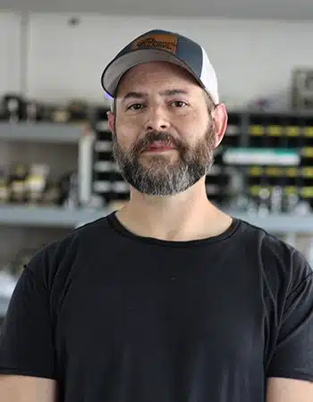 A bearded man wearing a black t-shirt and a baseball cap stands indoors with shelves of equipment in the background.