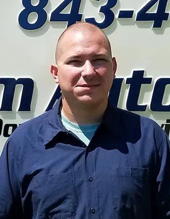 A man with a shaved head wearing a dark blue collared shirt stands in front of a sign with large blue and black text and numbers. He is looking at the camera and smiling slightly.