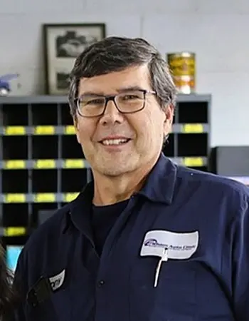 Kenny Moskalski, Founder/Owner Emeritus and 'Part-Time' staff member, smiles at the camera. He is wearing glasses and a dark blue button-up uniform shirt with 'Elham Auto Clinic' embroidered on it.