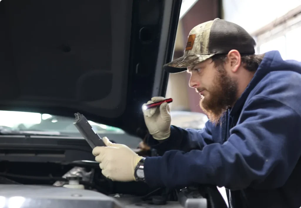 virginia state inspection, auto repair in West Point, VA at Eltham Auto Clinic. A mechanic wearing a cap and gloves examines a tablet while inspecting a car engine, holding a flashlight under the raised hood in a garage.
