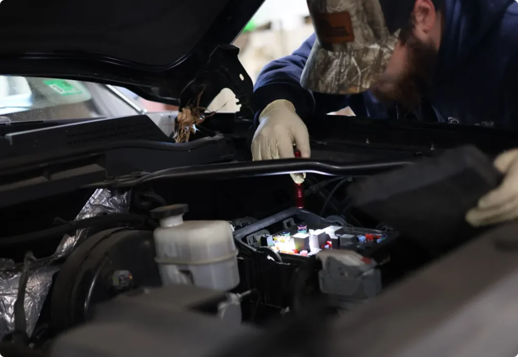 pre-purchase & pre-trip inspections. auto repair in West Point, VA at Eltham Auto Clinic. A mechanic wearing a camouflage hat and gloves works under the hood of a car, using a tool to inspect or repair the engine components.