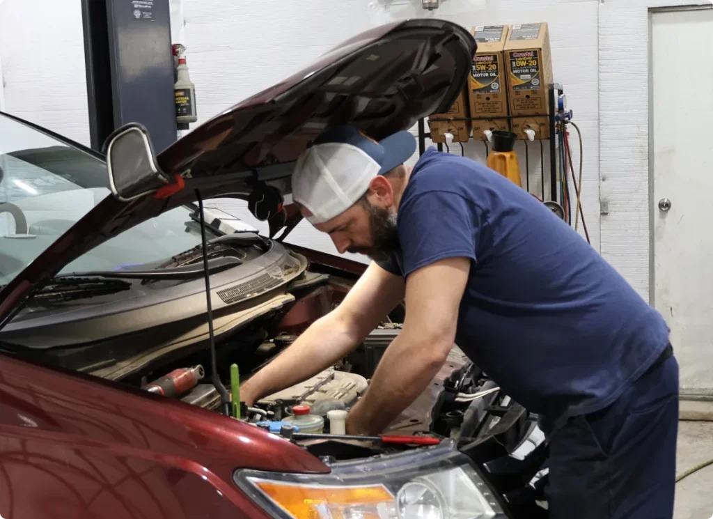 vehicle maintenance, auto repair in West Point, VA at Eltham Auto Clinic. A mechanic in a blue shirt and backward cap works under the hood of a red car in a garage, focusing on the engine. Tools and motor oil containers are visible in the background.