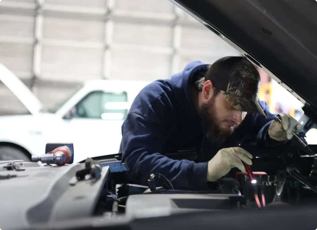 fleet maintenance, auto repair in West Point, VA at Eltham Auto Clinic. A bearded man wearing a cap, hoodie, and gloves works intently under the hood of a car in a garage. A white vehicle is visible in the background.