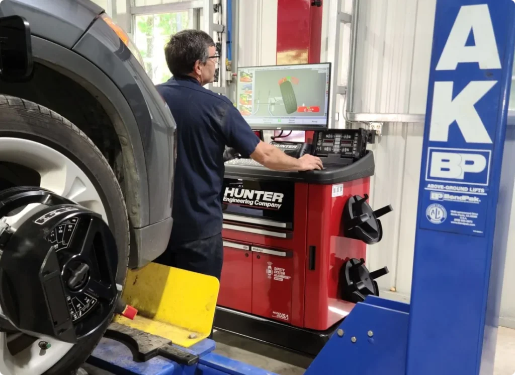 tire services, auto repair in West Point, VA at Eltham Auto Clinic. A mechanic uses a computer to check wheel alignment on a car in an auto repair shop. The vehicle’s front wheel is mounted on a lift with alignment equipment attached. Tools and a blue support beam are visible nearby.