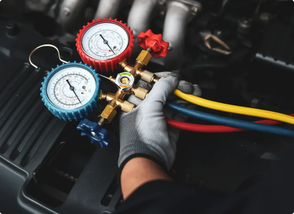 auto a/c & heater repair, auto repair in West Point, VA at Eltham Auto Clinic. A gloved hand holds a manifold gauge set with red, blue, and yellow hoses, used for diagnosing and servicing a cars air conditioning system, with part of an engine visible in the background.