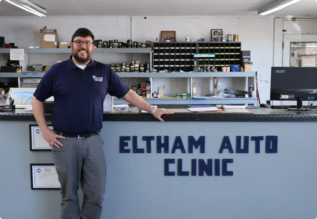 about us, auto repair in West Point, VA at Eltham Auto Clinic. A man in glasses and a navy shirt stands smiling at the counter of Eltham Auto Clinic, with tools and parts visible on shelves behind him inside the repair shop.