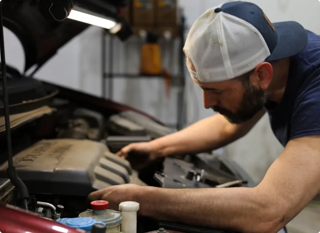 engine diagnostics, auto repair in West Point, VA at Eltham Auto Clinic. A man wearing a backward baseball cap works intently under the hood of a car, inspecting the engine in a garage setting.