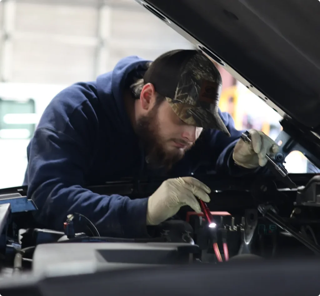 european, auto repair in West Point, VA at Eltham Auto Clinic. A mechanic wearing a camo cap, hoodie, and gloves works under the hood of a vehicle, using a light and tool to inspect or repair the engine.