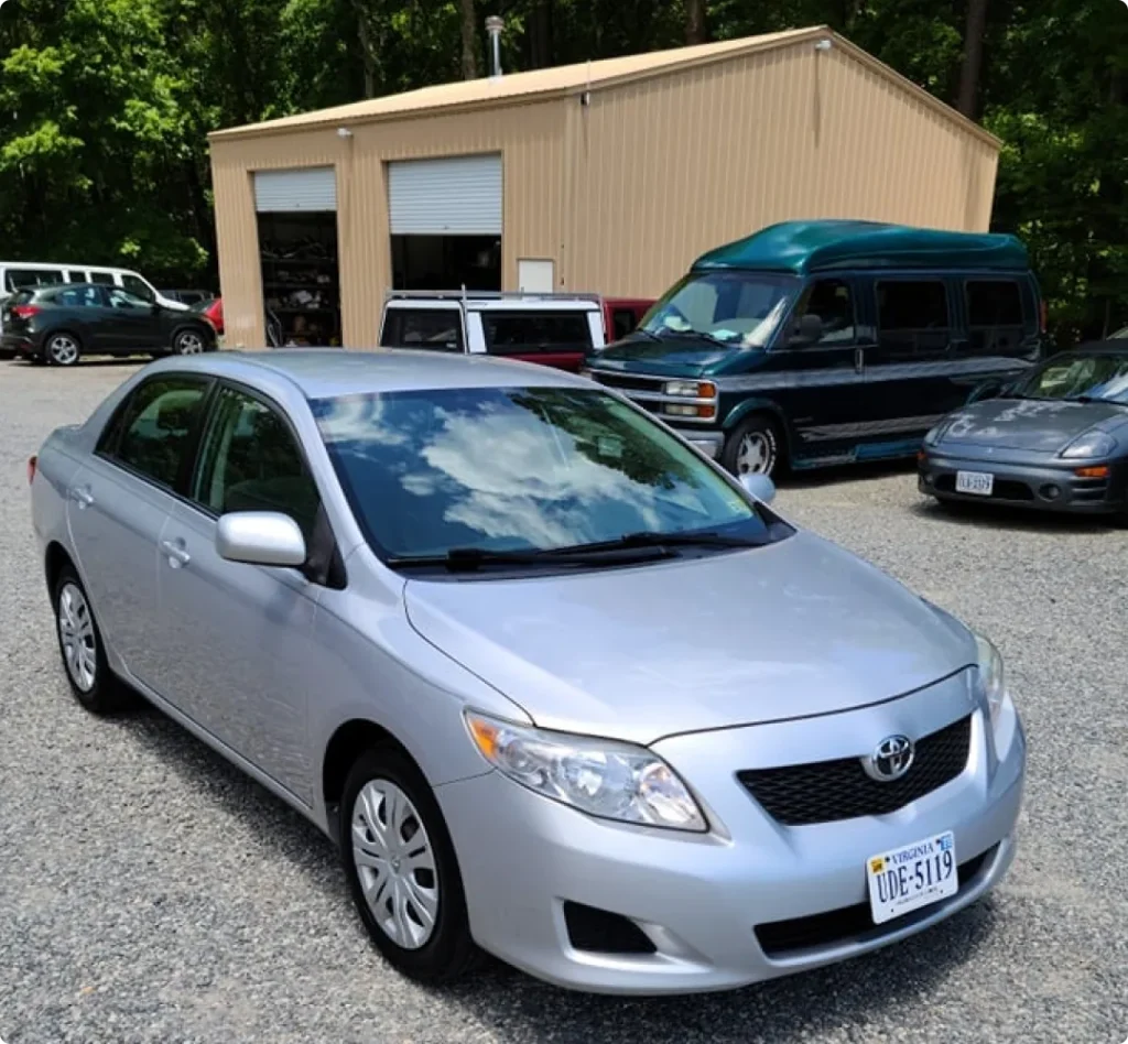 japanese, auto repair in West Point, VA at Eltham Auto Clinic. A silver Toyota sedan is parked on a gravel lot in front of a tan metal building, with other vehicles and trees visible in the background under a partly cloudy sky.
