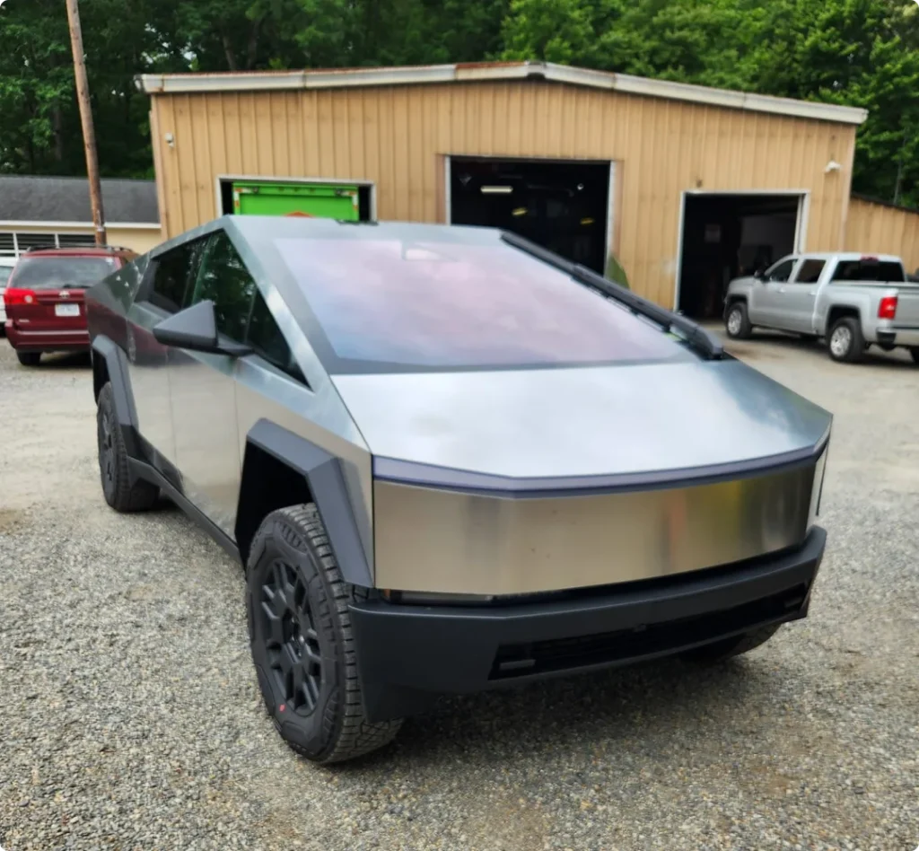 ev, auto repair in West Point, VA at Eltham Auto Clinic. A silver Tesla Cybertruck is parked on a gravel lot in front of a beige garage building, with trees and other vehicles in the background.
