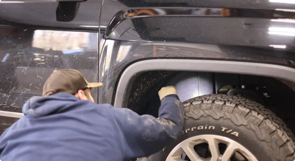 A person wearing a blue hoodie and cap inspects or works on the front wheel well of a black SUV, focusing on the tire and inner fender area.