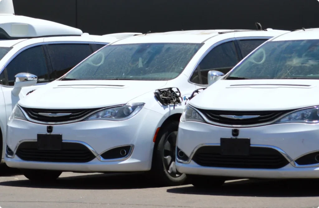 Several white minivans are parked in a row. One minivan has a missing side mirror and exposed wiring on the drivers side. The vehicles have no visible license plates.