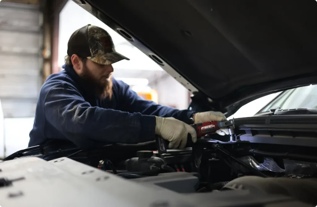 A mechanic wearing gloves and a camo hat uses a power tool to work under the hood of a car inside a garage.
