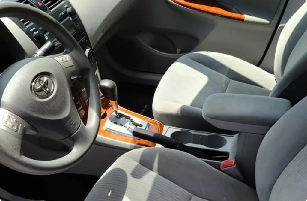 Interior of a car showing a grey fabric front seat, center armrest, gear shift, and steering wheel with the Toyota logo. Sunlight highlights the wood trim accents on the center console and door panel.