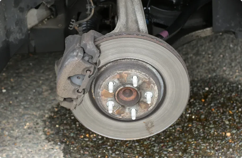 A close-up view of a car’s exposed brake disc and caliper, with the wheel removed. The metal surface shows signs of wear, and lug bolts are visible around the hub. The ground below is covered with small pebbles.
