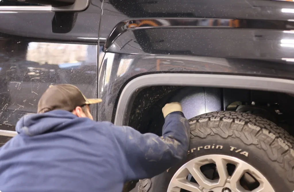 A person in a cap and hoodie inspects or works inside the wheel well of a black vehicle with mud-terrain tires.