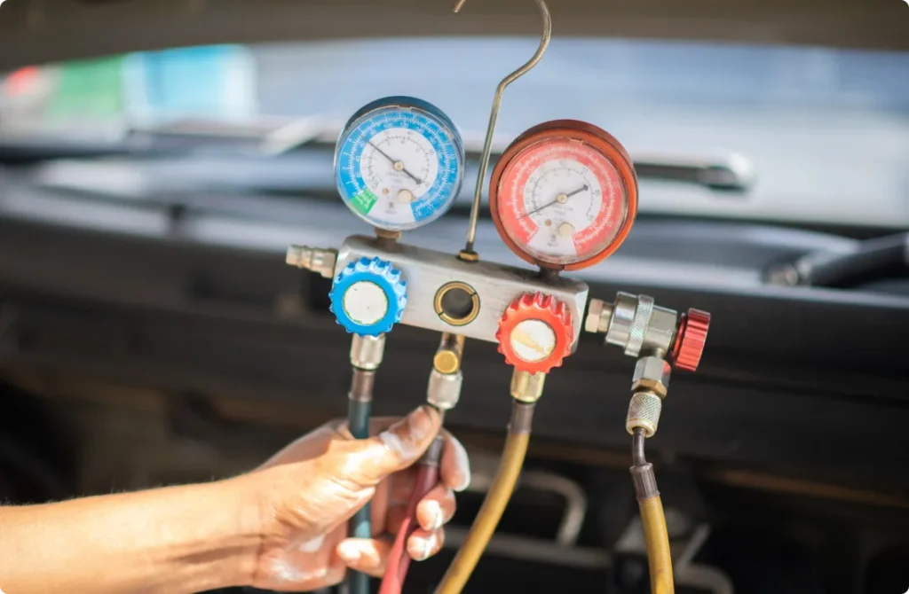 A hand holds a set of HVAC manifold gauges with blue and red dials connected to hoses, used to check and service a vehicle’s air conditioning system under its hood.