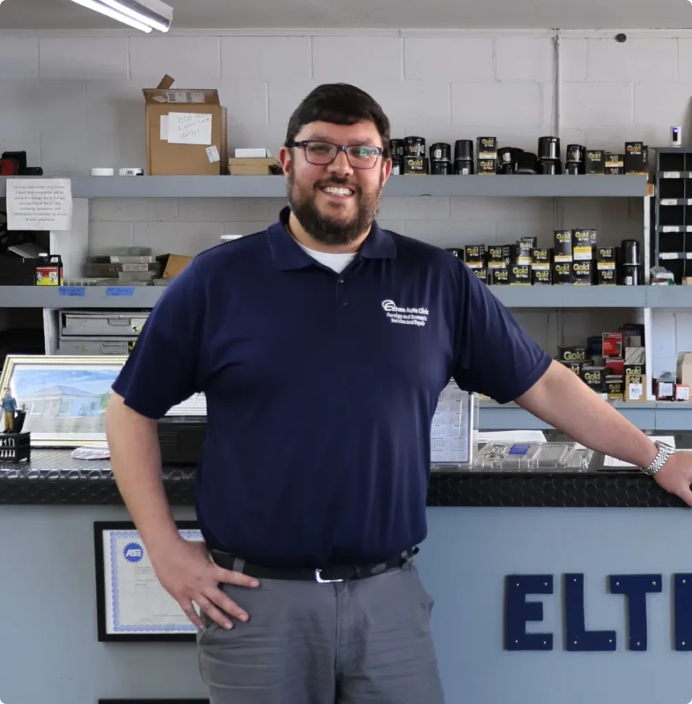 A man with glasses and a beard, wearing a navy polo shirt and gray pants, stands smiling with one hand on a counter in an auto shop, with shelves of car supplies and boxes in the background.