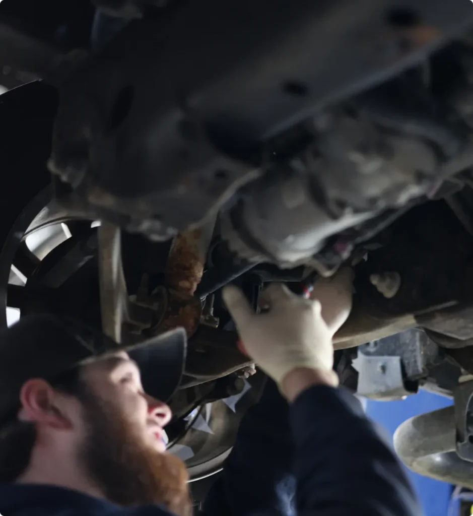 A mechanic wearing a cap and gloves works underneath a raised vehicle, focusing on the cars undercarriage components. The image is slightly blurred, emphasizing the mechanic’s face and hands.