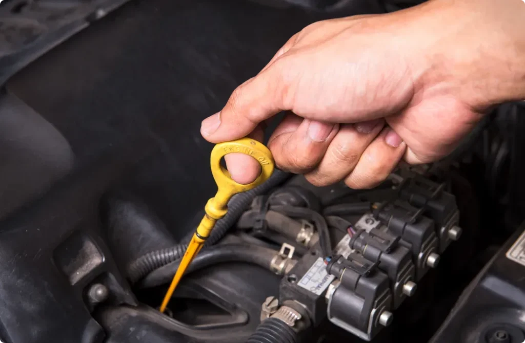 A hand pulls out a yellow engine oil dipstick from a car engine, checking the oil level. The engine components and wires are visible in the background.