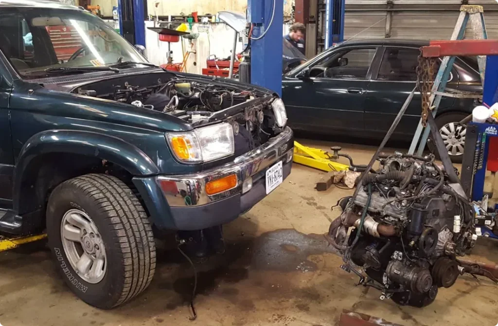 A dark green SUV with its hood open and engine removed sits in a garage. The engine lies on the floor nearby, and another black sedan is parked in the background amid shop equipment.