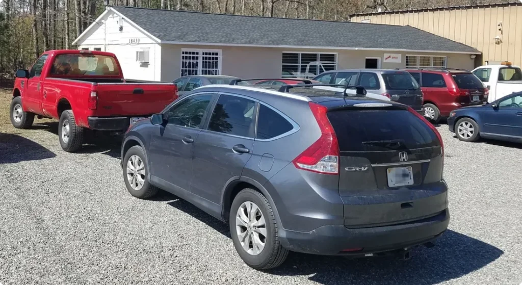 A gray Honda CR-V and a red pickup truck are parked on a gravel lot in front of a white building with several windows and other vehicles visible in the background.