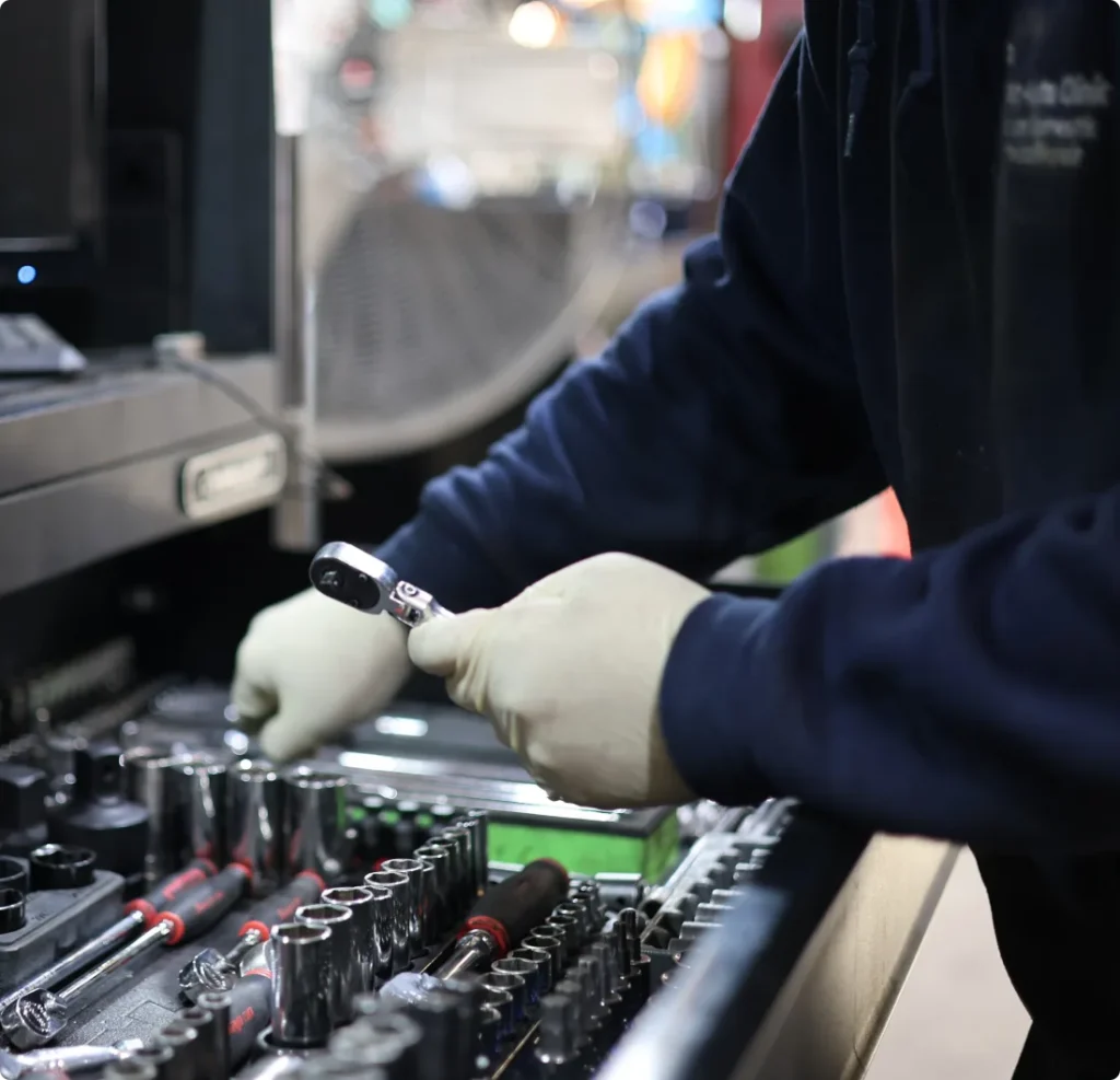 A person wearing light-colored gloves organizes or selects a socket wrench from a drawer filled with various tools and sockets in a workshop or garage setting.